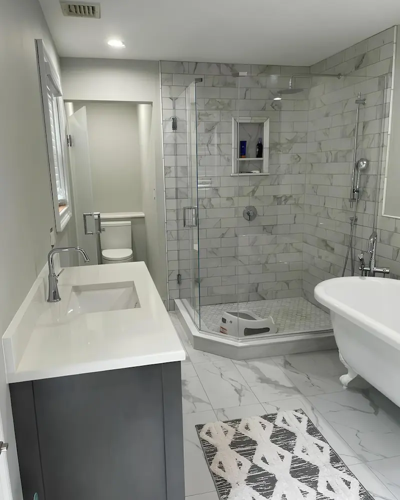 Transitional primary bathroom with heritage clawfoot soaking tub, neo-angle frameless marble subway tile shower enclosure with hex-mosaic floor, and dark charcoal vanity with integrated white solid-surface top in Pelham Manor, Westchester County — JMR Construction